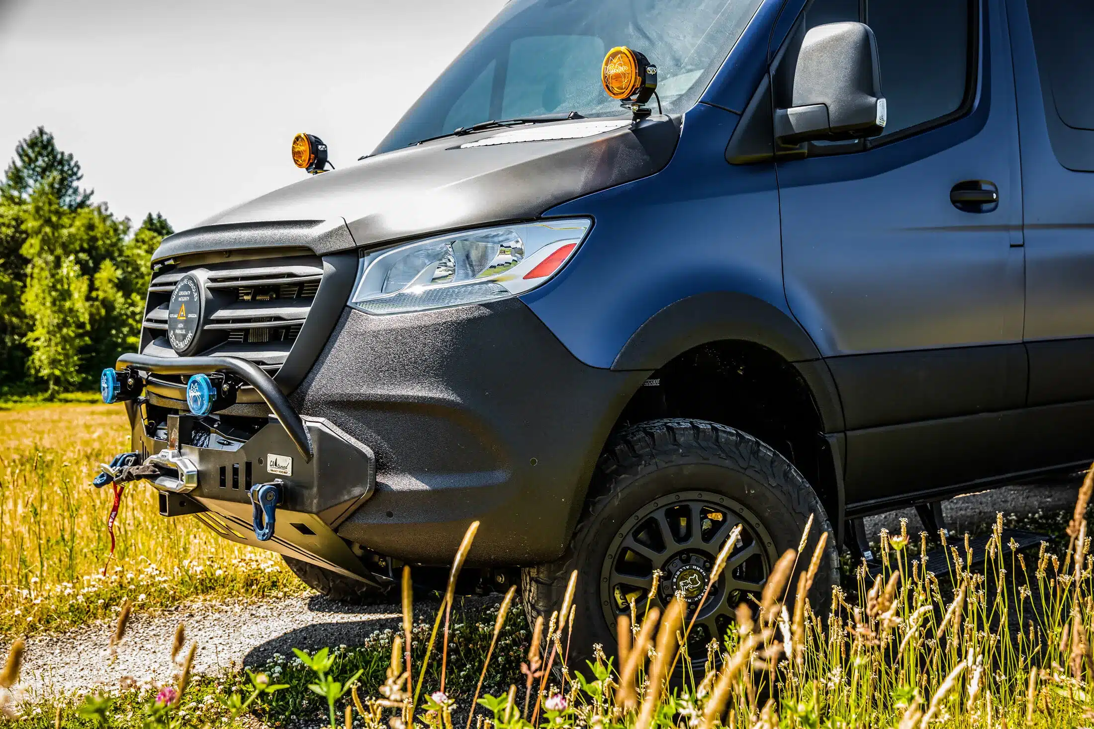 Nahaufnahme der Front eines Mercedes Sprinter 170 2500 4x4 Campervans mit Offroad-Stoßstange, Zusatzscheinwerfern und grobstolligen Reifen auf einer Wiese