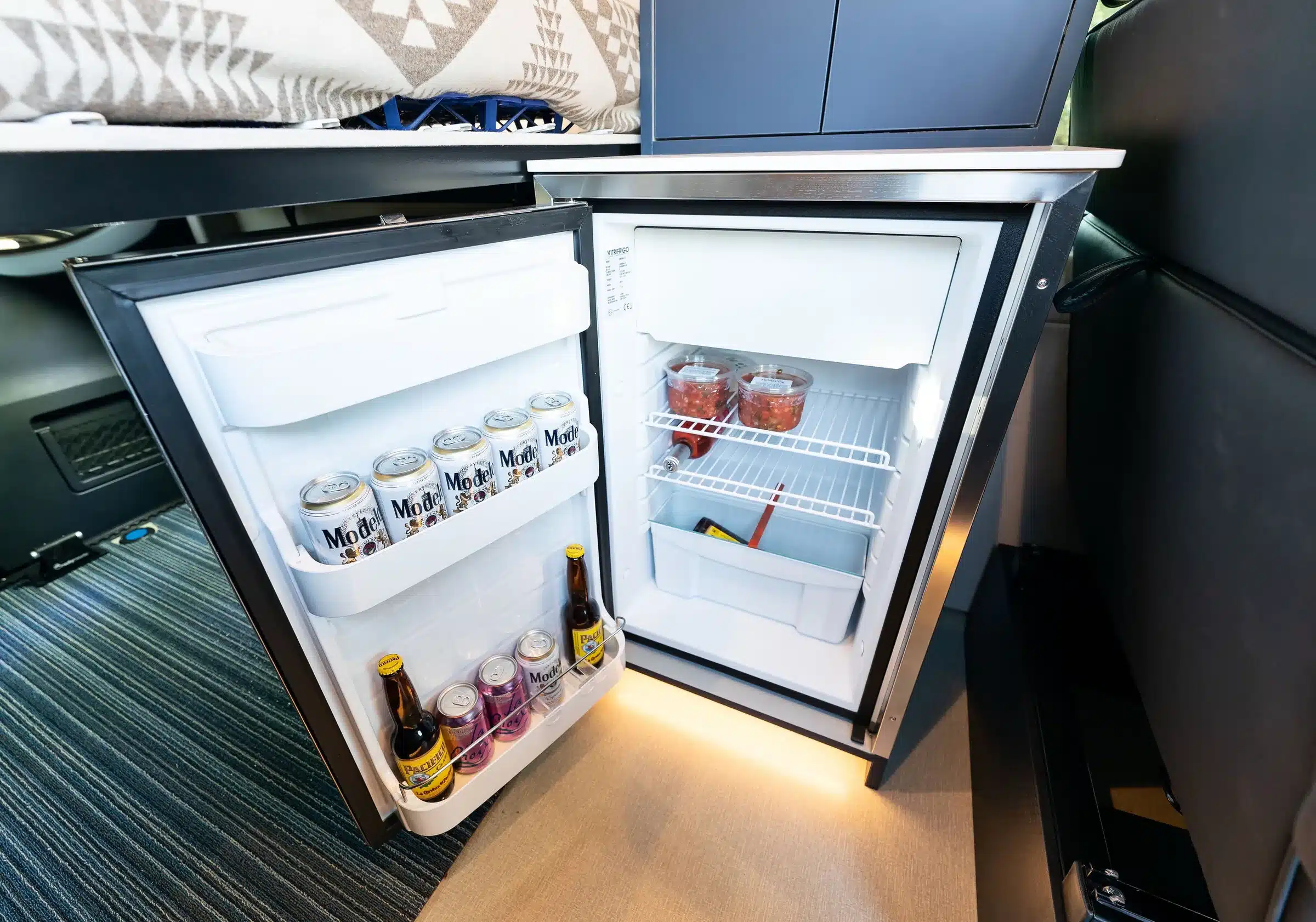 Offener Kühlschrank mit Getränkedosen, Flaschen und Lebensmitteln im Innenraum eines ausgebauten Mercedes Sprinter Vans
