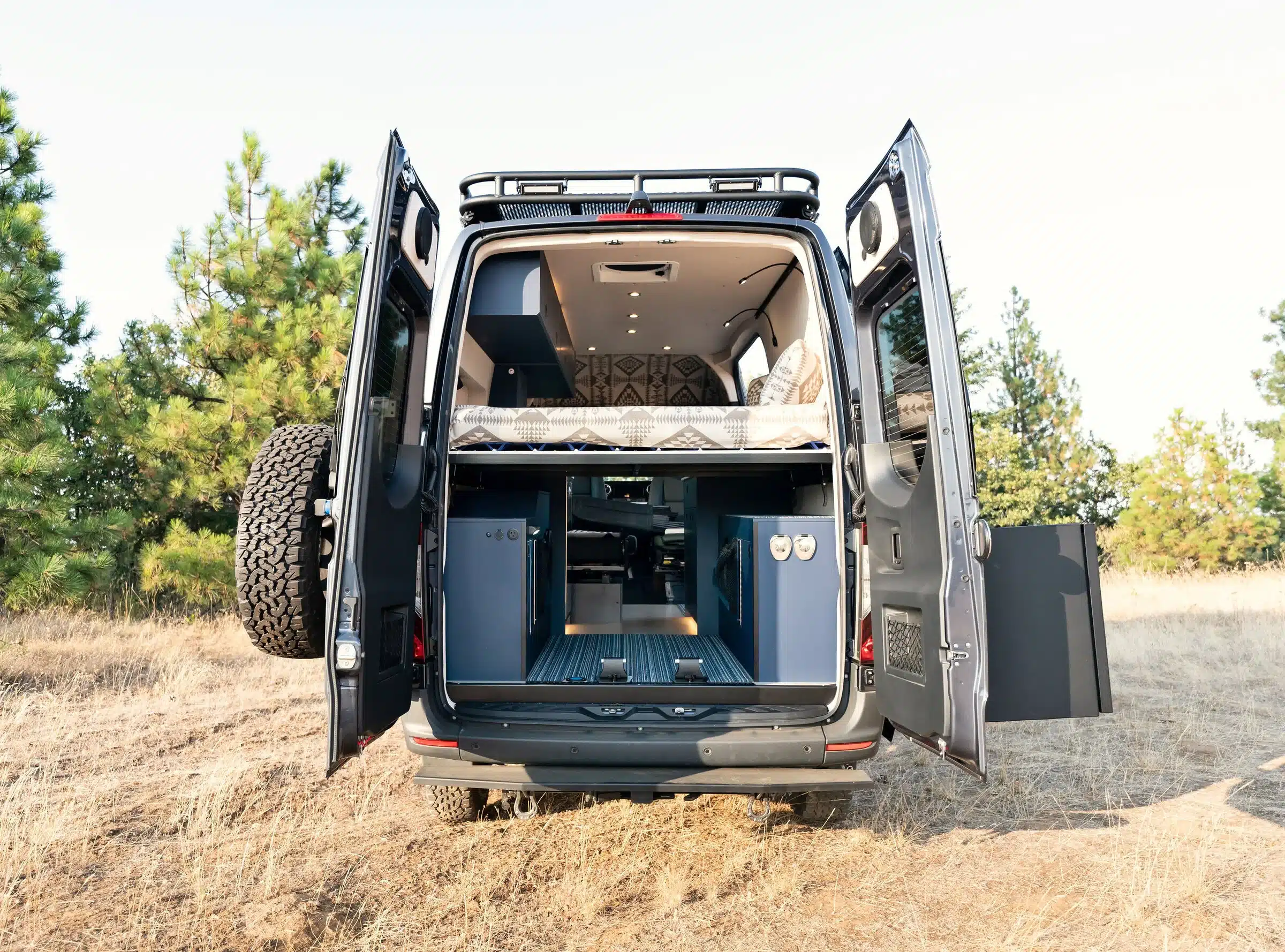 Geöffnete Hecktüren eines ausgebauten Mercedes Sprinter 4x4 mit Plattformbett, Stauraum und Camper-Innenausstattung.