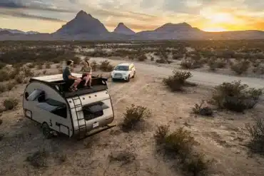 Alto F1743 Expedition Wohnanhänger mit zwei Personen auf dem Dach, abseits der Straße in wüstenartiger Landschaft, im Hintergrund ein SUV und Berge bei Sonnenuntergang