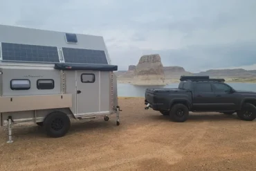 AntiShanty AS-1 A-Frame Trailer mit Solarpanels, angehängt an einem schwarzen Pickup, steht auf sandigem Untergrund vor einem See und Felsenlandschaft