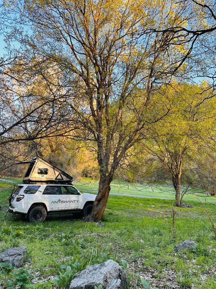 Geländewagen mit aufgeklapptem, hartwandigem Dachzelt-Aufsatz (Roof Top Dwelling) von Antishanty, geparkt unter Bäumen auf einer Wiese