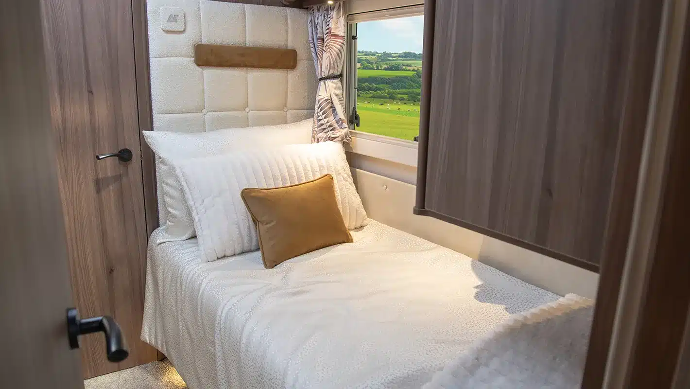 Einzelbett mit heller Bettwäsche und Kissen im Innenraum eines Auto-Sleepers Burford Duo Wohnmobils, daneben ein Fenster mit Blick ins Grüne