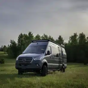 bashie 4x4-Sprinter Campervan steht auf einer grünen Wiese vor einem Wald, mit Dachträger und Offroad-Ausstattung