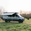 Mercedes Sprinter 170 4x4 von Benchmark Vehicles mit ausgefahrener Markise auf einer Wiese, umgeben von Bäumen