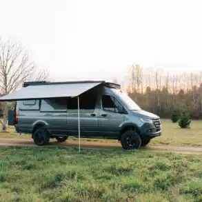Mercedes Sprinter 170 4x4 von Benchmark Vehicles mit ausgefahrener Markise auf einer Wiese, umgeben von Bäumen