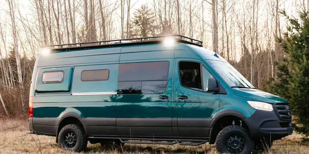 Grüner Benchmark Acorn 4x4 Sprinter Campervan mit Offroad-Bereifung und Dachträger steht auf einer Wiese vor Bäumen