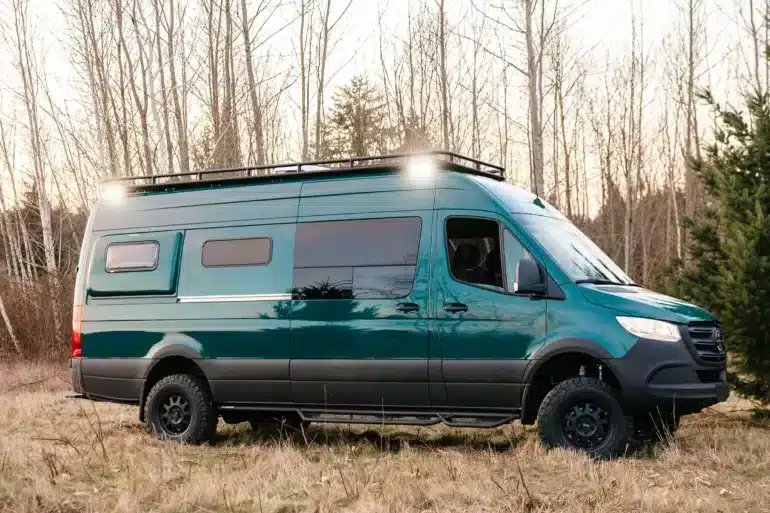 Grüner Benchmark Acorn 4x4 Sprinter Campervan mit Offroad-Bereifung und Dachträger steht auf einer Wiese vor Bäumen