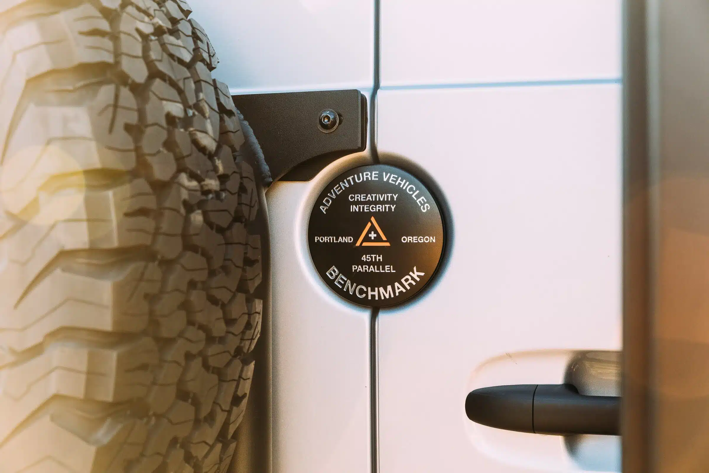 Detailaufnahme eines Benchmark Adventure Vehicles Emblems neben einem grobstolligen Reserverad an einem weißen Mercedes Sprinter