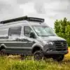 Grauer Benchmark Elkat Expeditions-Van mit Offroad-Ausstattung steht auf einer Wiese vor bewölktem Himmel.