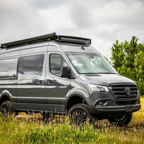 Grauer Benchmark Elkat Expeditions-Van mit Offroad-Ausstattung steht auf einer Wiese vor bewölktem Himmel.