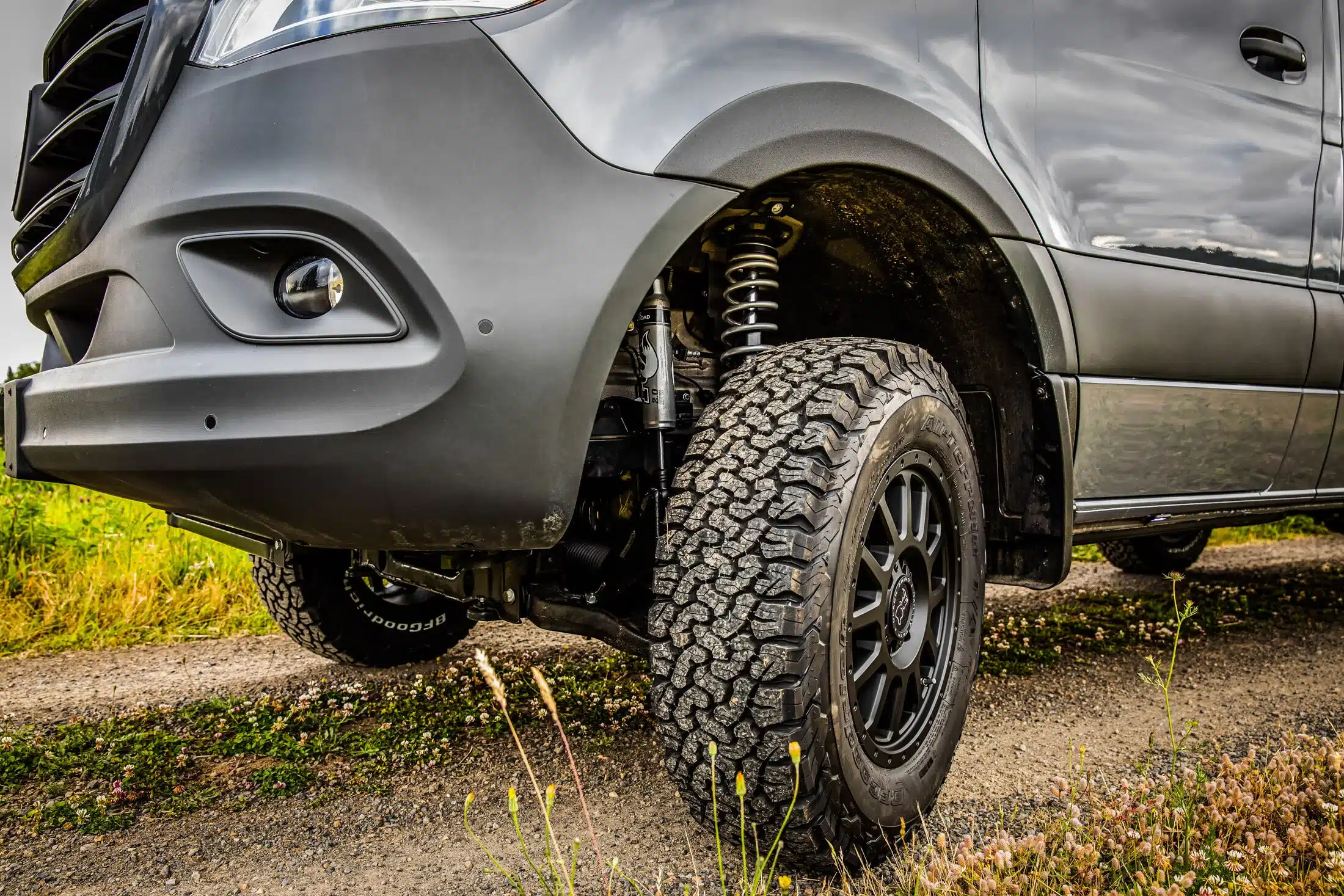 Nahaufnahme eines Offroad-Camper-Vans mit grobstolligen Geländereifen und verstärktem Fahrwerk auf einem Feldweg