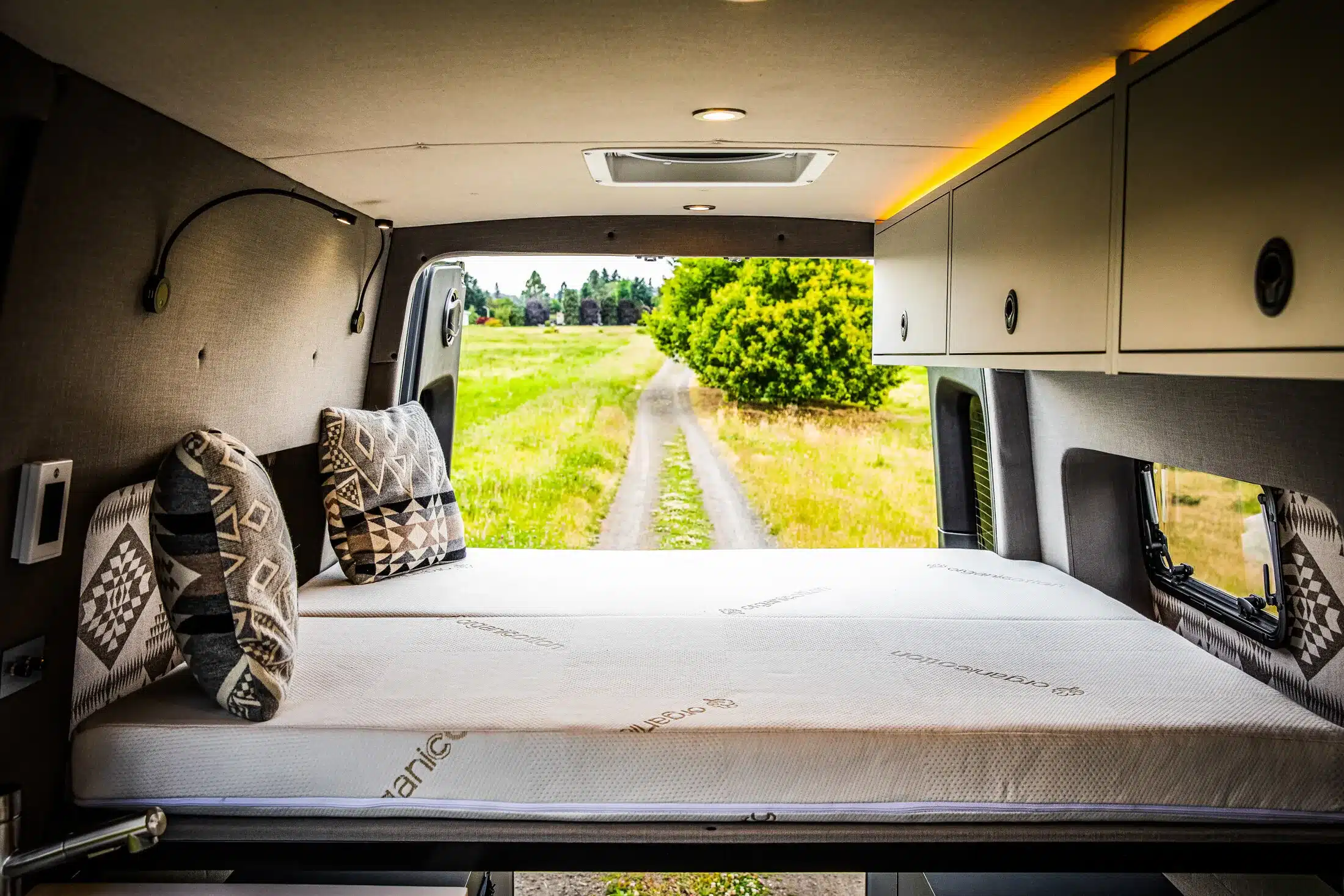 Blick aus dem Schlafbereich eines ausgebauten Camper-Vans Benchmark Elkat auf einen Feldweg und grüne Landschaft.