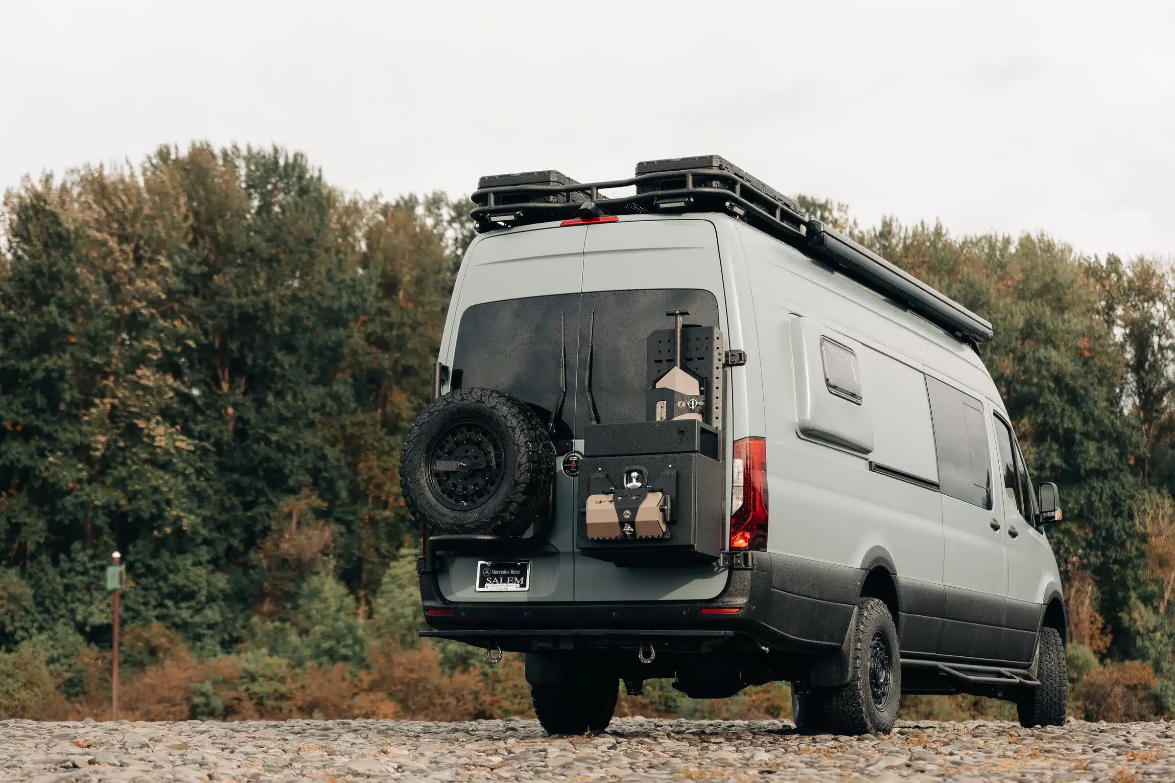 Heckansicht eines ausgebauten Mercedes Sprinter 170 EXT AWD mit Offroad-Komponenten und Dachträger auf einem Schotterplatz