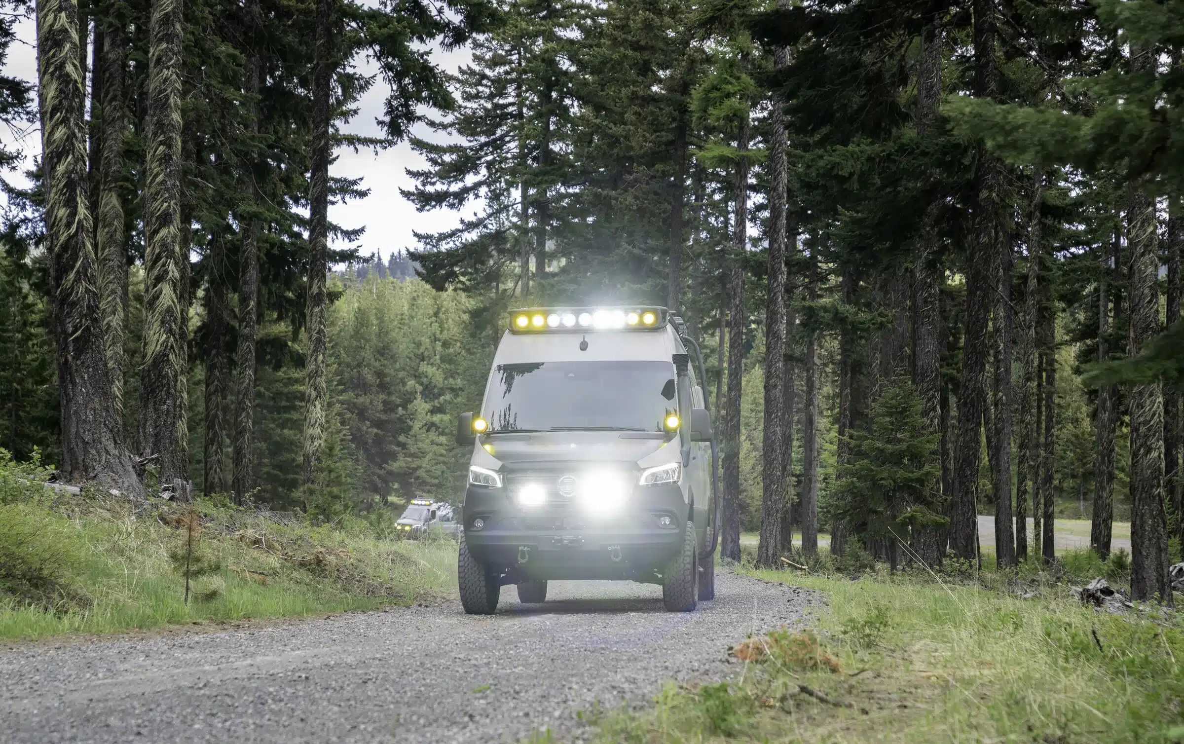 Mercedes Sprinter 4x4 Benchmark Mojave mit Dachträger und Zusatzbeleuchtung fährt auf einem Waldweg
