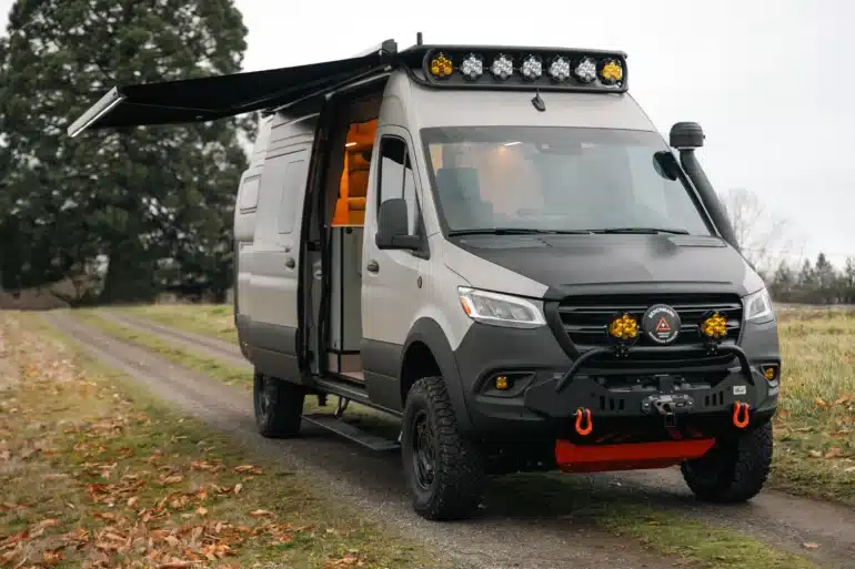 Benchmark Revive Sprinter 2024 Campervan mit Offroad-Ausstattung, Markise und geöffneter Seitentür auf Feldweg