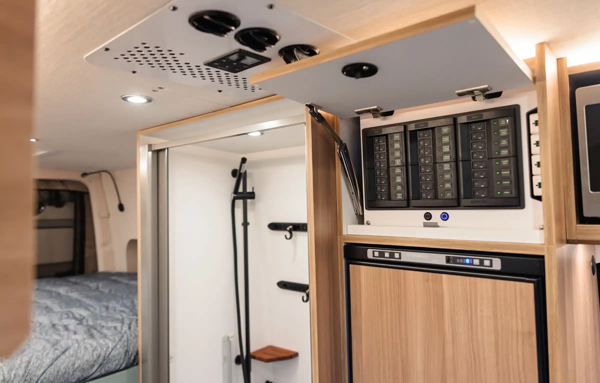 Innenansicht eines Benchmark Revive Sprinter Vans mit geöffneter Elektro-Schaltzentrale, Duschkabine und Vitifrigio Slim 150 Kühlschrank