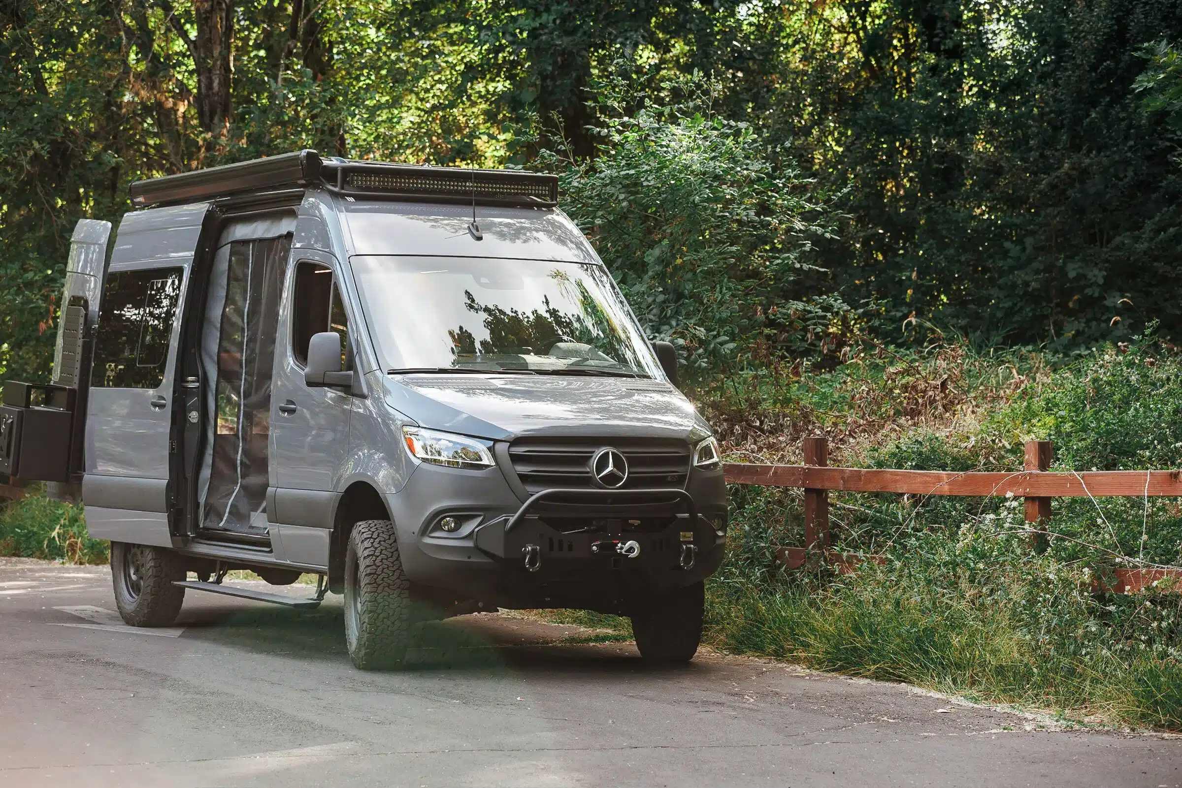 Grauer Benchmark Rule 76 Adventure Van mit Offroad-Ausstattung steht am Waldrand auf einer Straße, Schiebetür geöffnet