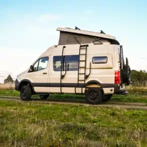Seitenansicht eines beigen Mercedes Sprinter 144 4x4 Campervans mit Aufstelldach und Leiter, geparkt auf einem Feldweg