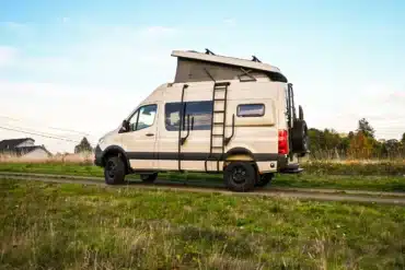 Seitenansicht eines beigen Mercedes Sprinter 144 4x4 Campervans mit Aufstelldach und Leiter, geparkt auf einem Feldweg