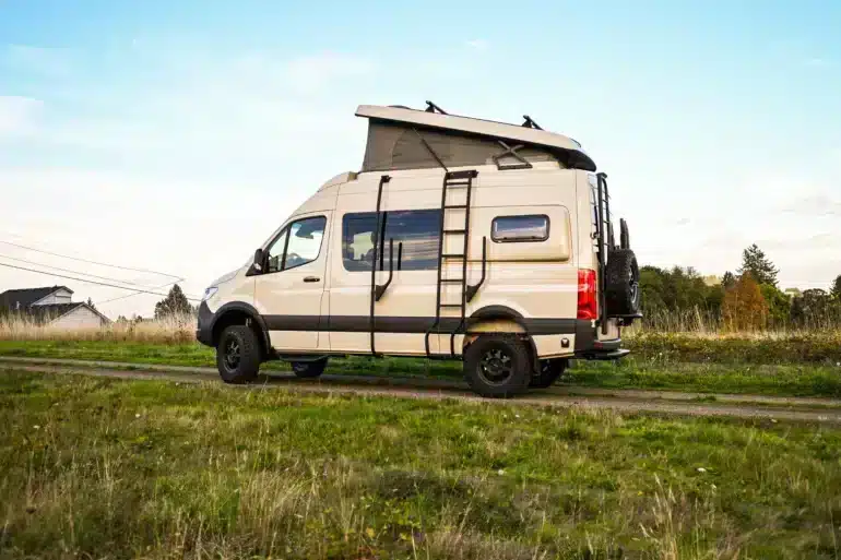 Seitenansicht eines beigen Mercedes Sprinter 144 4x4 Campervans mit Aufstelldach und Leiter, geparkt auf einem Feldweg
