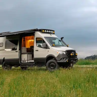 Ausgebauter Benchmark Vehicles Cortland Campervan mit Markise, geöffneten Türen und Offroad-Ausstattung auf einer grünen Wiese