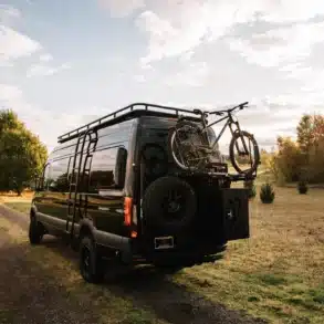 Schwarzer Mercedes-Benz Sprinter 170 4x4 Campervan mit Fahrradträger und Ersatzrad am Heck, geparkt auf einem Feldweg bei Sonnenuntergang