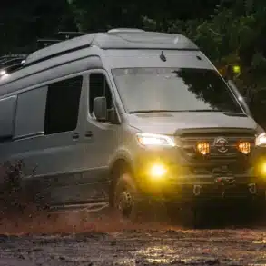 Big Sur Sprinter 4x4 Camper fährt durch schlammiges Gelände im Wald, Scheinwerfer leuchten