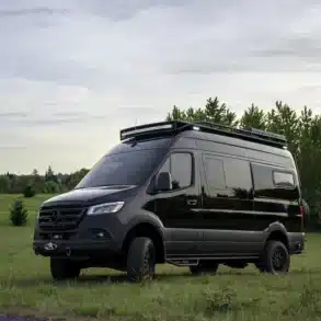 Schwarzer Mercedes Sprinter 144 HR 4x4 Campervan mit Dachträger und Offroad-Ausstattung auf einer Wiese