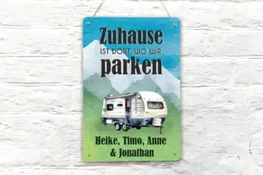 Buntes Blechschild mit Wohnwagen und dem Spruch 'Zuhause ist dort, wo wir parken', personalisiert mit Namen, vor weißer Wand