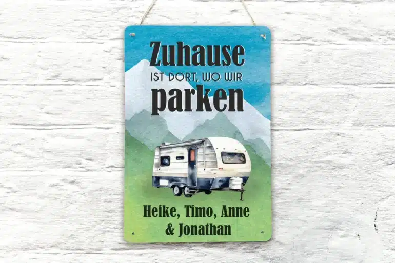 Buntes Blechschild mit Wohnwagen und dem Spruch 'Zuhause ist dort, wo wir parken', personalisiert mit Namen, vor weißer Wand
