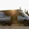 Seitenansicht eines ausgebauten Mercedes Sprinter 170 4x4 Campervans mit geöffneter Schiebetür und Markise, aufgenommen im Sonnenuntergang am Flussufer