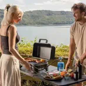 Zwei Personen grillen mit dem Campingaz Tour & Grill CG an einem Seeufer, der kompakte Gasgrill steht auf einem Campingtisch mit Grillgut und Zutaten.