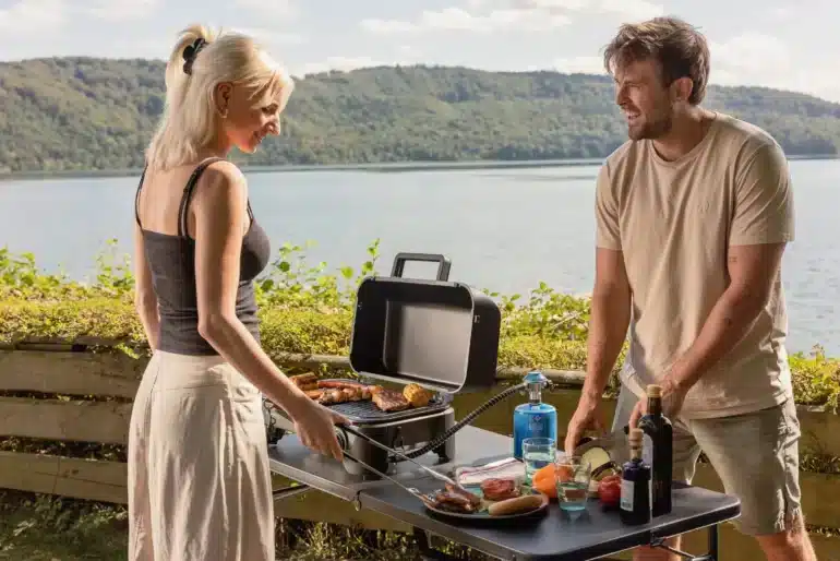 Zwei Personen grillen mit dem Campingaz Tour & Grill CG an einem Seeufer, der kompakte Gasgrill steht auf einem Campingtisch mit Grillgut und Zutaten.