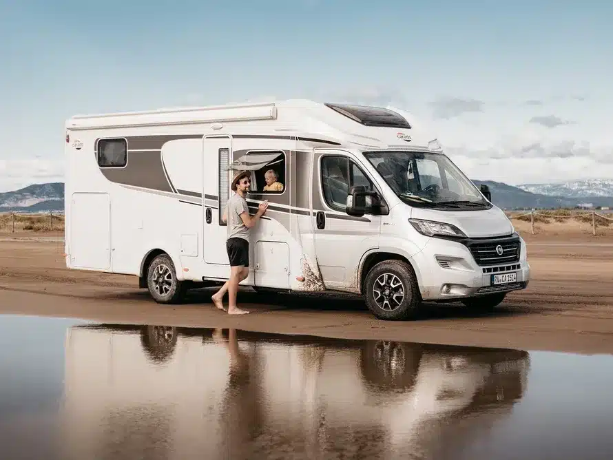Carado T448 teilintegriertes Wohnmobil mit Personen am Fenster und Eingang, geparkt am Strand mit Spiegelung im Wasser