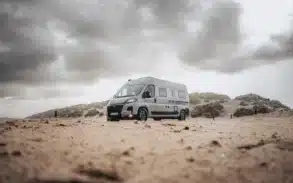 CROSSCAMP ADVTR 6.4 EF Kastenwagen-Camper steht auf sandigem Untergrund vor bewölktem Himmel