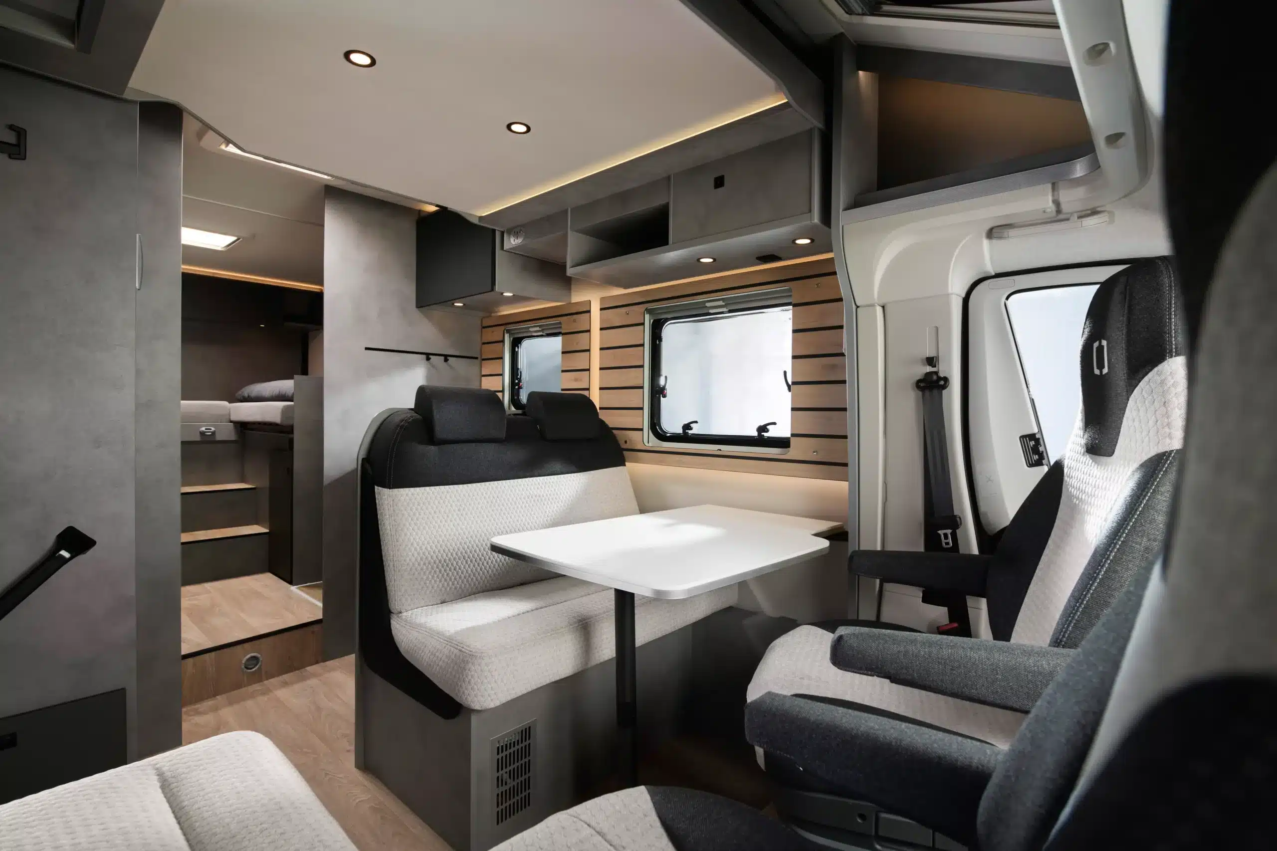 Innenansicht des Crosscamp EXPDN 7.4 E mit Sitzgruppe, Tisch, drehbaren Fahrersitzen und Blick auf die Einzelbetten im Heck