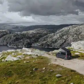 Ein Crosscamp EXPLR Campervan mit geöffnetem Aufstelldach steht auf einem Parkplatz an einem See, umgeben von felsiger, grüner Berglandschaft und bewölktem Himmel