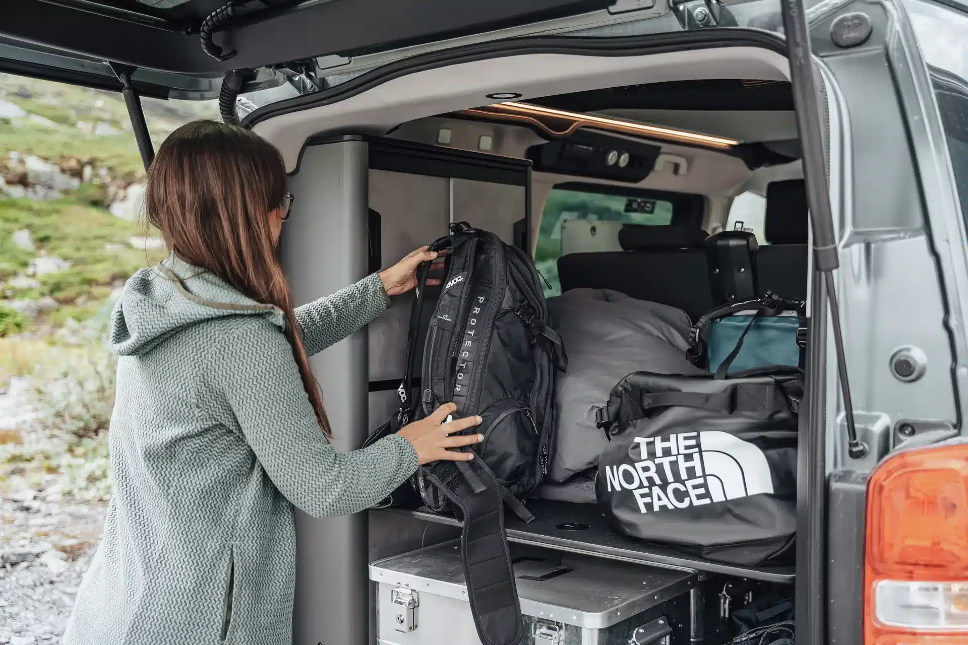Frau verstaut einen Rucksack im Kofferraum eines Crosscamp EXPLR Campervans, umgeben von Taschen und Campingausrüstung