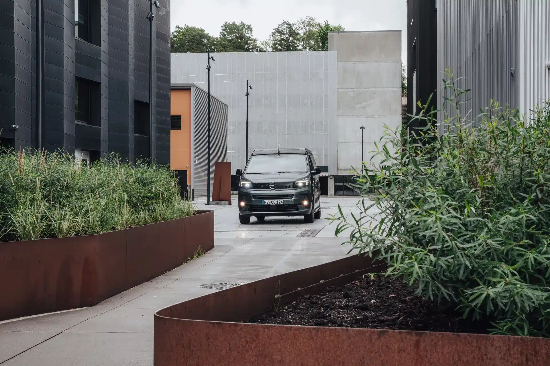 Ein Crosscamp EXPLR Campervan fährt durch eine moderne Stadtlandschaft mit Gebäuden und Pflanzenbeeten