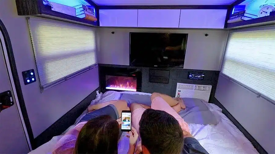 Paar liegt im Bett des inTech Luna Teardrop Trailers, Blick auf Fernseher, Kamin und Stauraum