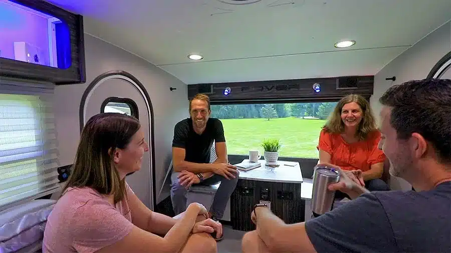Vier Personen sitzen im Innenraum eines inTech Luna Teardrop Trailers und unterhalten sich an einem Tisch, große Fenster bieten Blick ins Grüne.