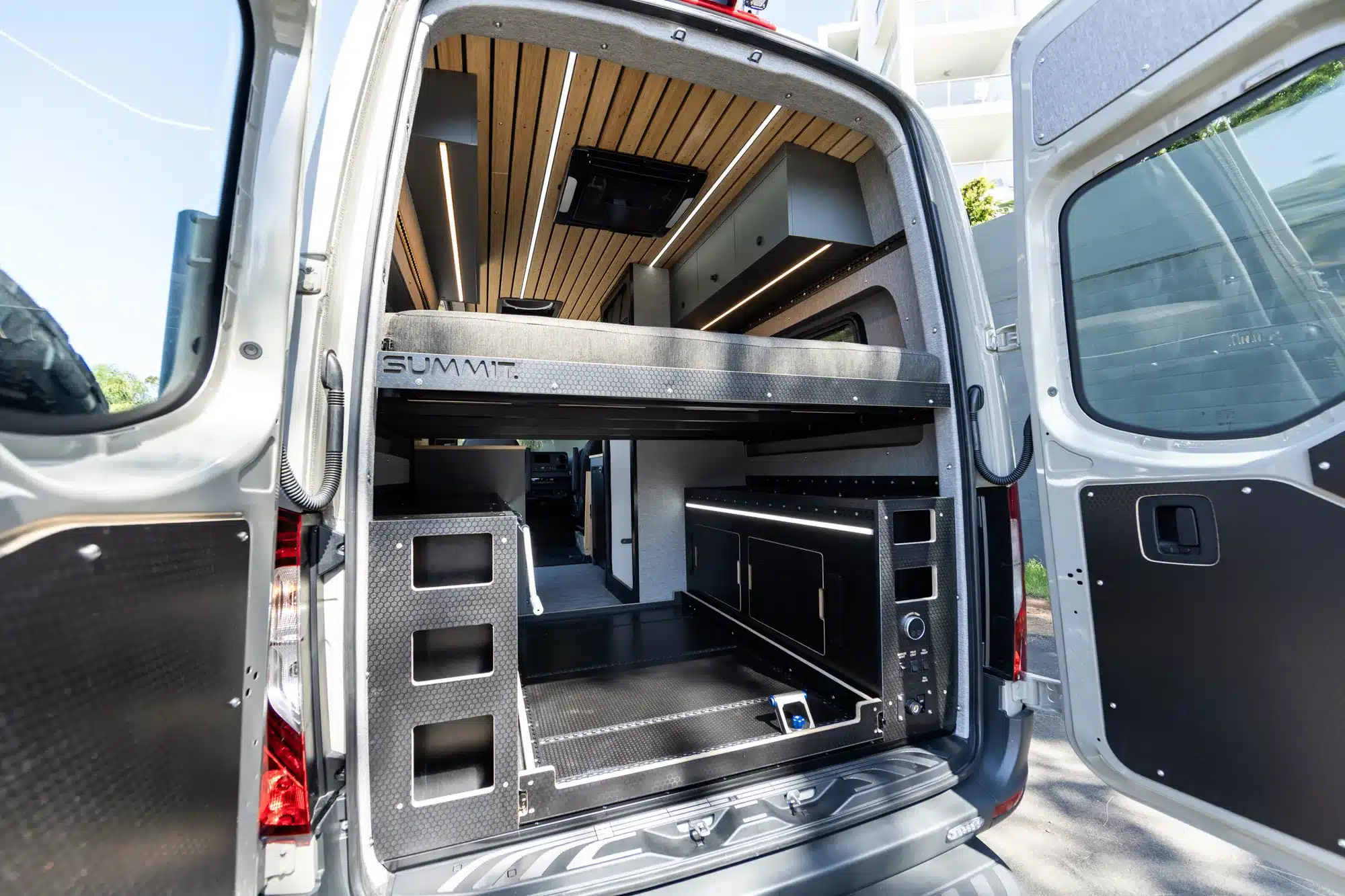 Offene Hecktüren eines Summit M1 Vans mit sichtbarem Stauraum, Bettpodest und hochwertigem Innenausbau