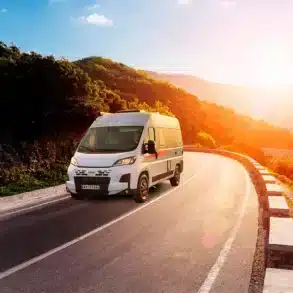 Weißer Kastenwagen V540G fährt auf einer kurvigen Straße durch eine sonnige Landschaft mit Bergen im Hintergrund.