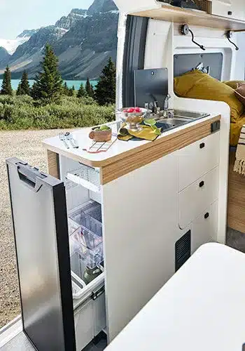 Offene Camper-Küche im V540G mit herausgezogenem Kühlschrank, Spüle, Schubladen und Ausblick auf Berge und See