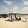 Zwei Personen sitzen unter einem aufblasbaren Dometic HUB 2 REDUX Pavillon am Strand neben einem Offroad-Fahrzeug und Campingausrüstung.