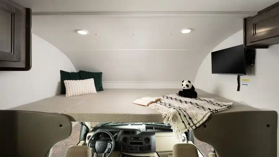 Innenansicht des Alkovenbetts über dem Fahrerhaus im Entegra Coach Esteem XL mit Kissen, Decke, Buch und Fernseher