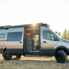 Seitenansicht eines silbernen Eskadi 4x4 Sprinter Vans mit geöffnetem Einstieg, Offroad-Bereifung und Dachträger, stehend auf einer Wiese bei Sonnenuntergang