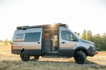 Seitenansicht eines silbernen Eskadi 4x4 Sprinter Vans mit geöffnetem Einstieg, Offroad-Bereifung und Dachträger, stehend auf einer Wiese bei Sonnenuntergang