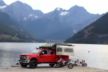 Roter Pick-up mit Far Out Canopy Truck Camper und zwei Motorrädern am Seeufer vor Bergkulisse